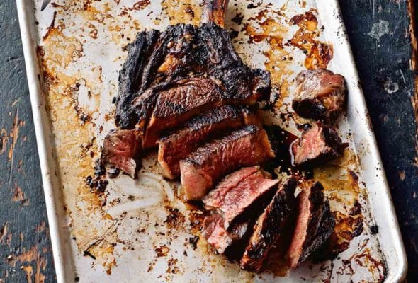 A sliced Korean steak on a rimmed baking sheet with a knife resting on the sheet and bowls of kimchi and daikon nearby.