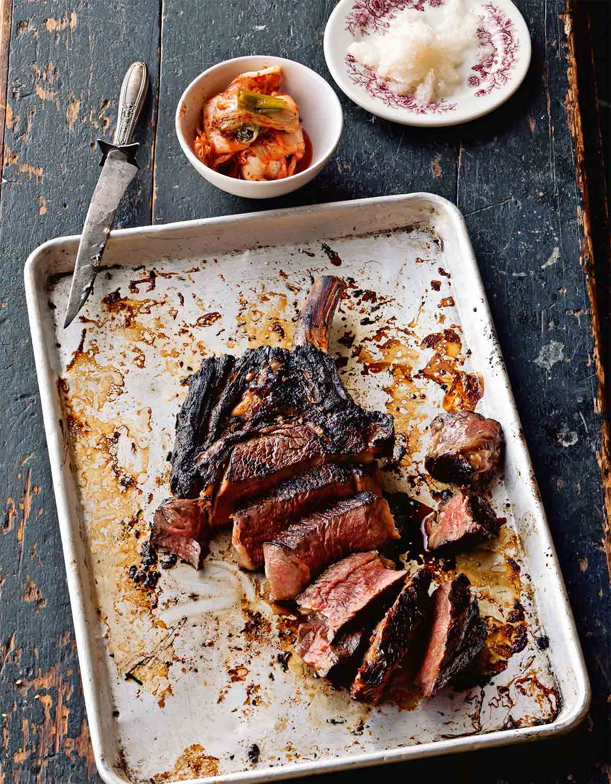 A sliced Korean steak on a rimmed baking sheet with a knife resting on the sheet and bowls of kimchi and daikon nearby.