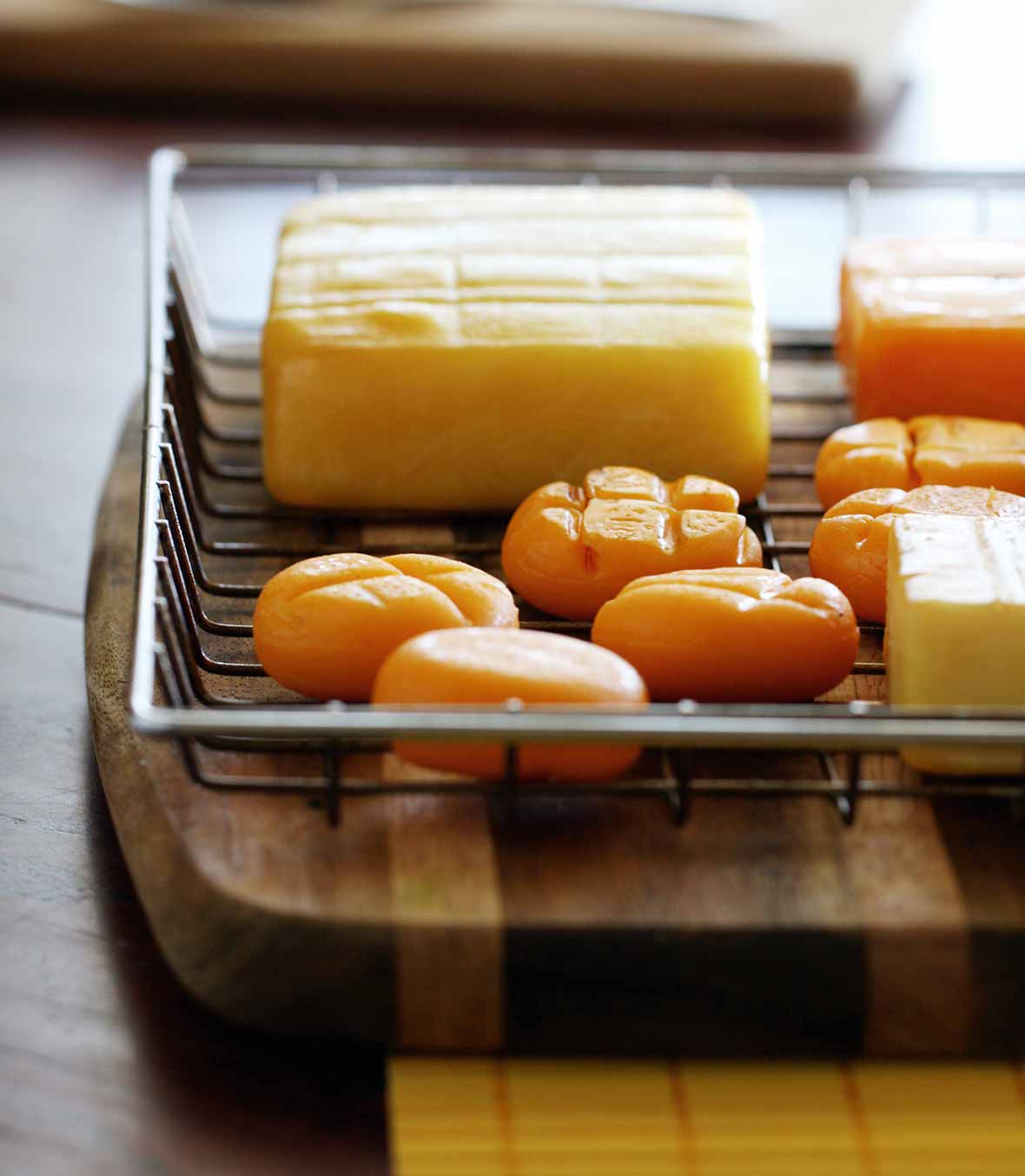 A wire basket on a wood cutting board containing several types of cheese, including smoked cheddar cheese