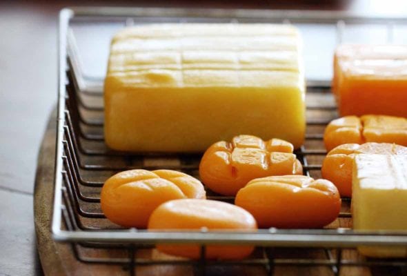 A wire basket on a wood cutting board containing several types of cheese, including smoked cheddar cheese
