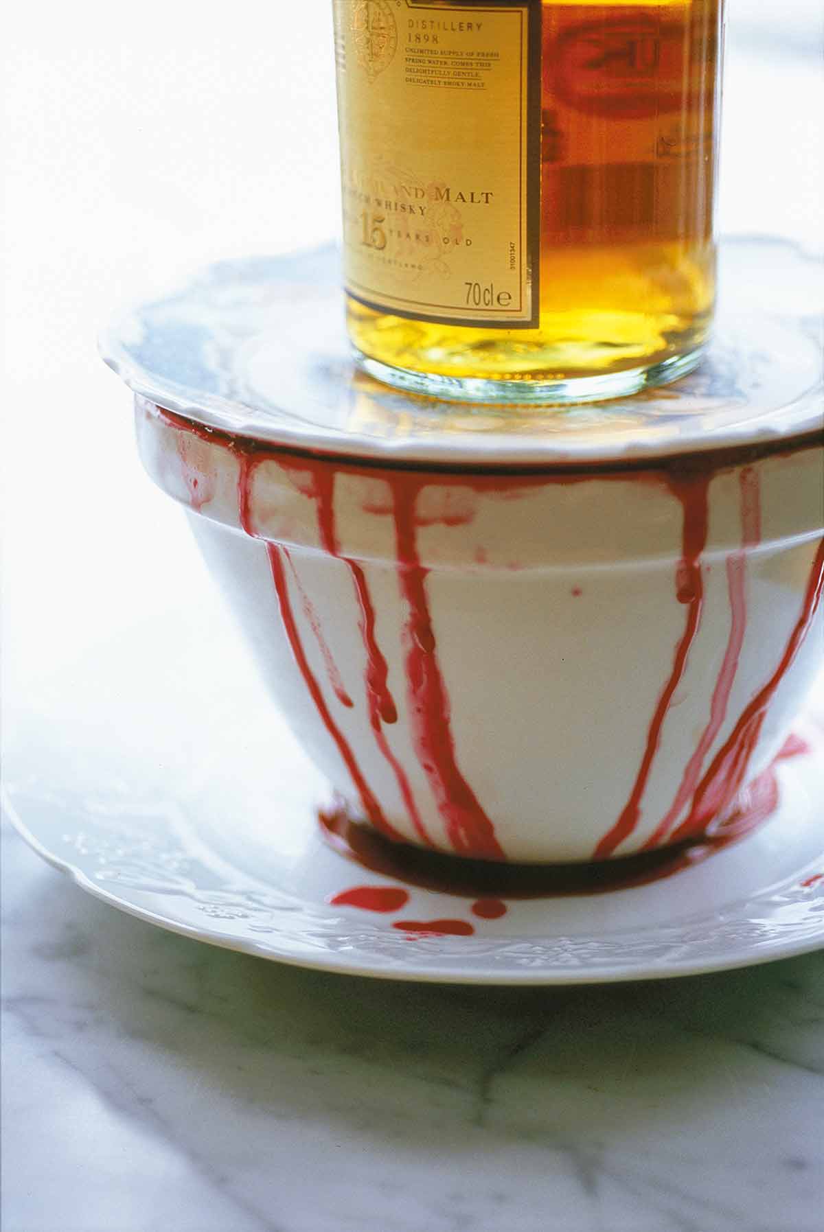 A white bowl of wine-soaked autumn pudding with wine dripping down the side topped with a white plate and a bottle of whiskey.