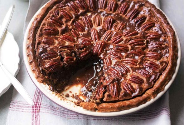 A chocolate pecan pie in a ceramic pie plate with one slice missing on a linen napkin.