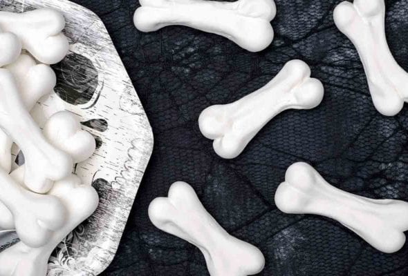 Halloween meringue bones piled on a paper plate and scattered on a black cobweb tablecloth beside the plate.