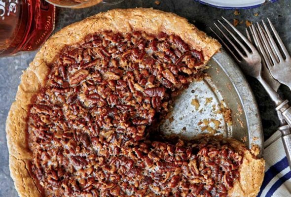 A maple pecan pie in a metal pie plate with one slice missing and forks, plates, and a bottle of rye whiskey beside the pie.