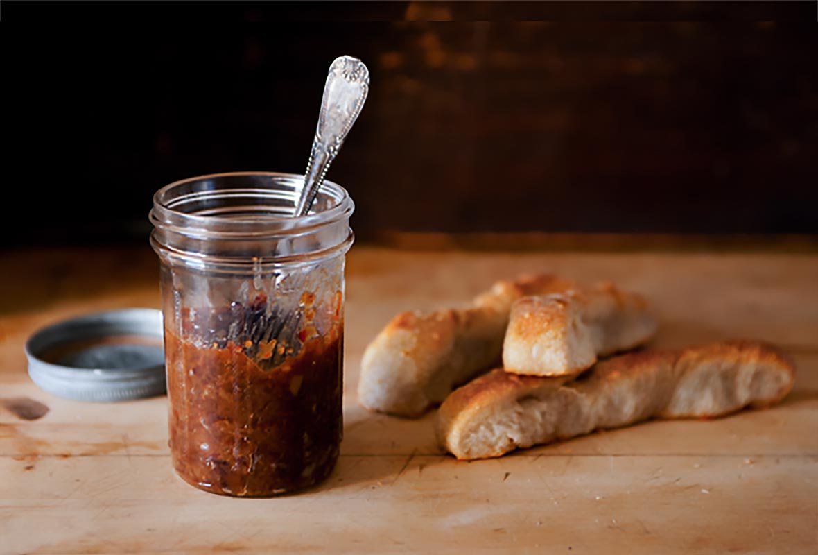 A small Mason jar half filled with bacon jam on a wooden board with a few pieces of bread beside the jar.