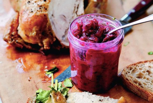 An open jar of cranberry chutney on a cutting board next to a partially carved chicken and a sandwich being assembled.
