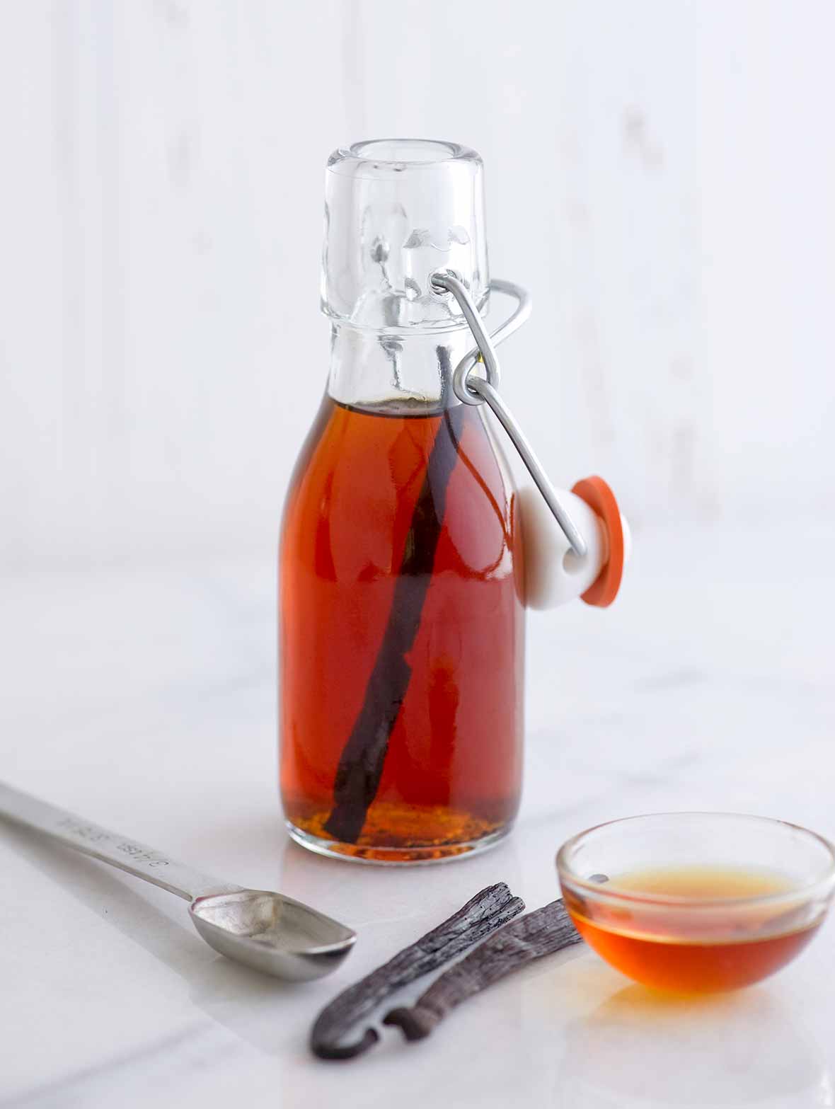 A small bottle of homemade vanilla extract, a measuring spoon, two vanilla beans, and a small bowl of amber liquid.