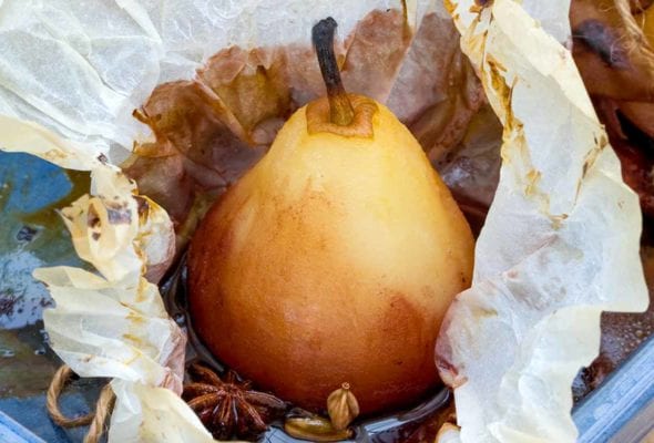 A whole peeled pear in an opened piece of parchment with star anise and cardamom in a blue baking dish.