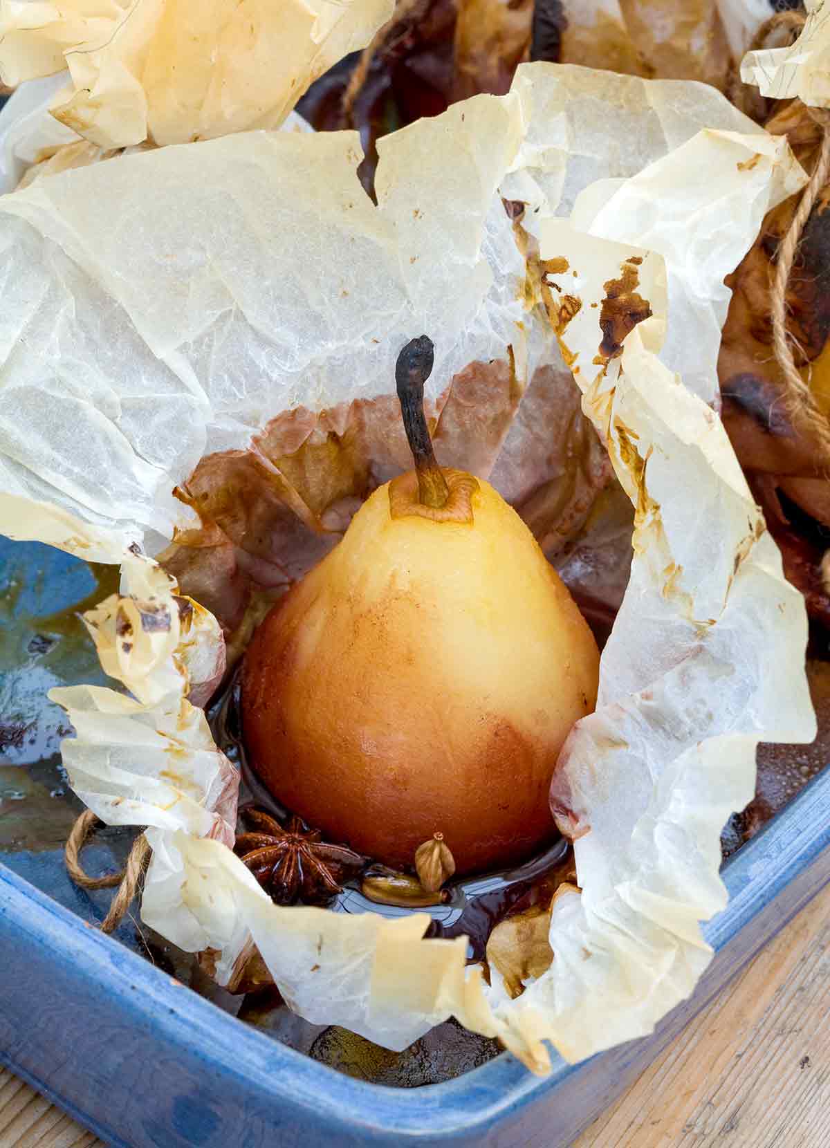 A whole peeled pear in an opened piece of parchment with star anise and cardamom in a blue baking dish.