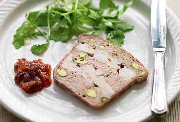 A white plate topped with a slice of chicken, pork, and pistachio terrine, a dollop of chutney, some arugula leaves and a knife.