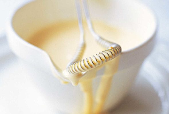 A dish of classic Hollandaise sauce on a white plate with a whisk resting on the dish.