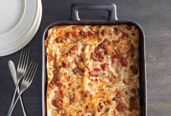 A metal casserole dish filled with a cooked lasagna Bolognese, with three forks and a stack of plates beside it.