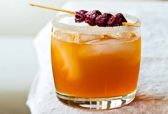 A highball glass with a sugared rim is filled with sidecar cocktail, ice cubes and a dried cherry garnish.