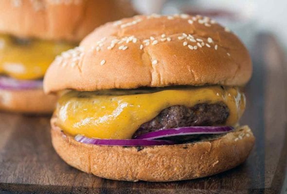 Two classic cheeseburgers with red onion and pickles on sesame buns on a wooden serving board.
