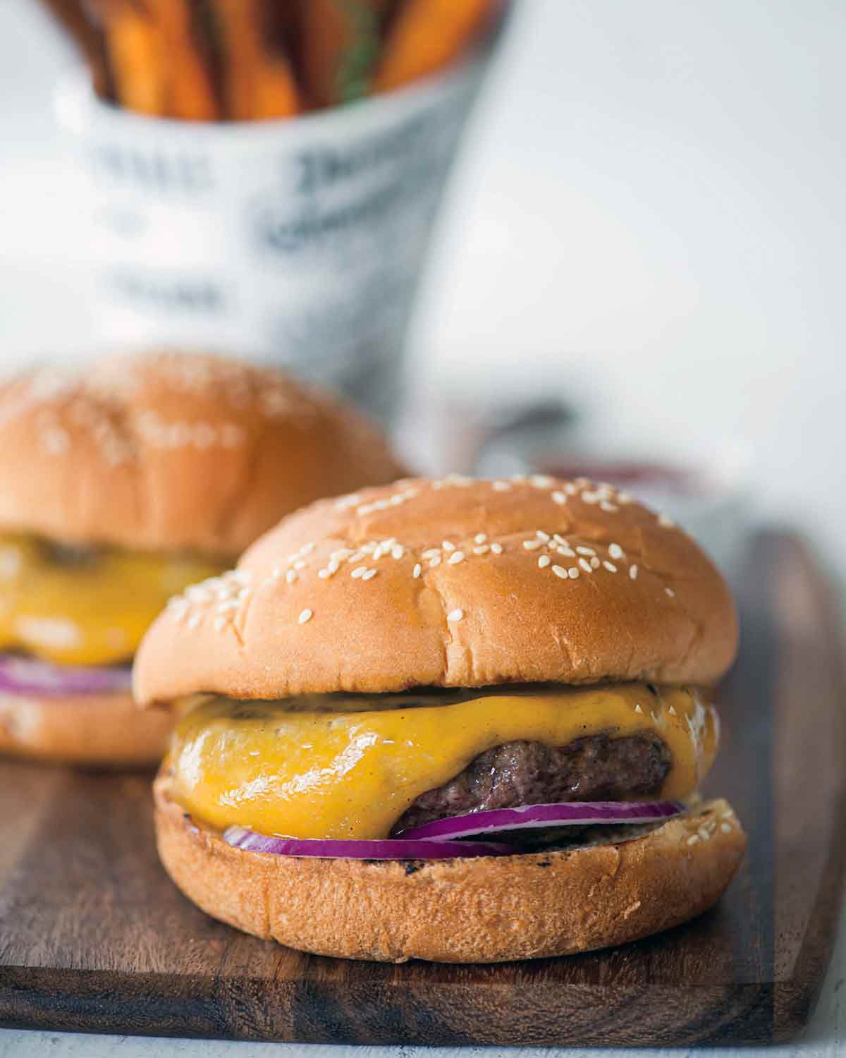 Two classic cheeseburgers with red onion and pickles on sesame buns on a wooden serving board.