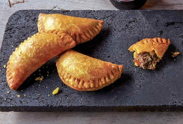 Five half-moon pastries, Jamaican beef turnovers, on a stone plate, three bottles of beer
