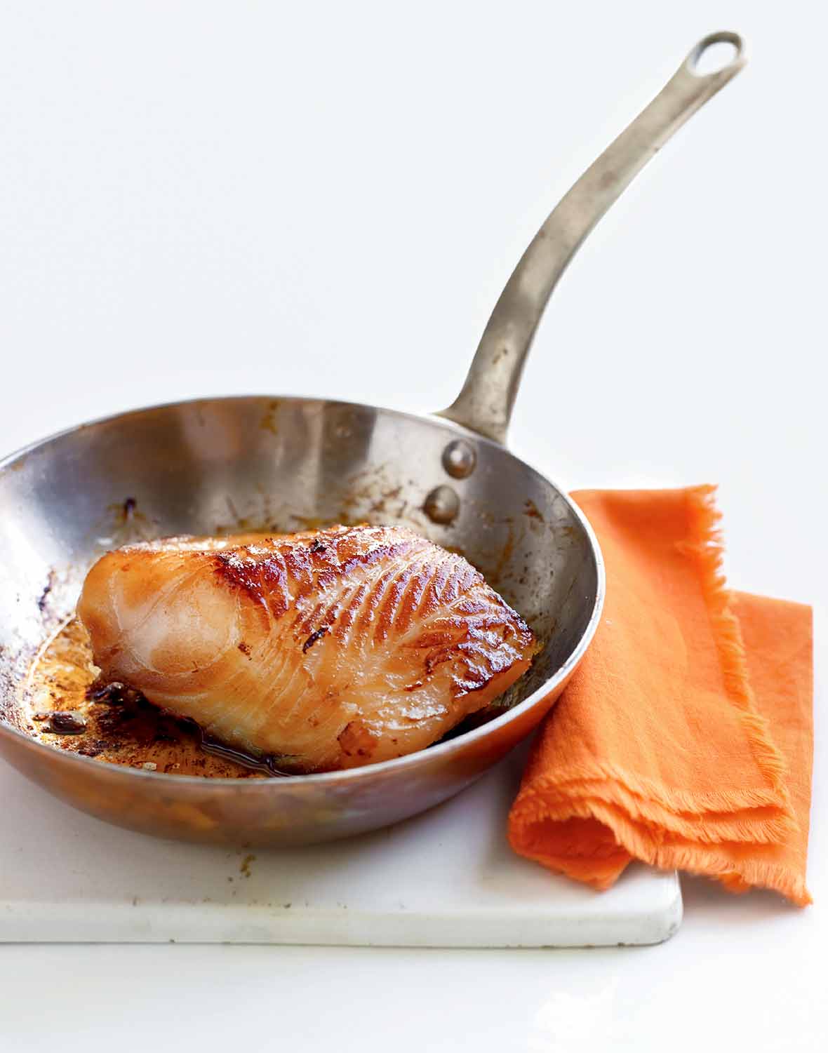 A piece of cooked miso cod in a stainless steel skillet that is resting on a white board next to an orange napkin.