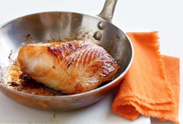 A piece of cooked miso cod in a stainless steel skillet that is resting on a white board next to an orange napkin.