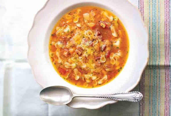 A white bowl filled with pasta soup with potatoes and pancetta on a linen cloth with a spoon resting on the edge of the bowl.