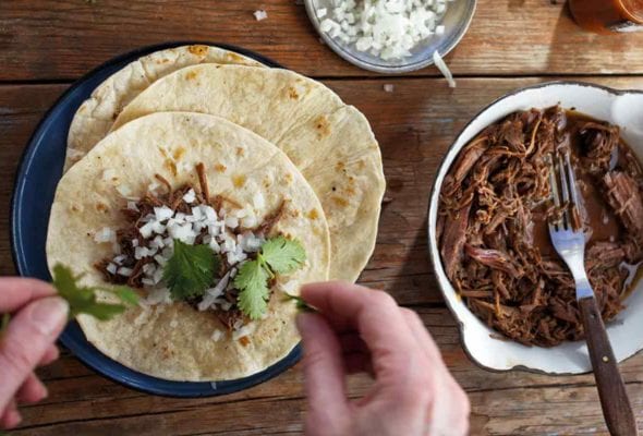 A person assembling a slow cooker barbacoa taco with a pot of shredded barbacoa meat, a small dish of onions, a bottle of hot sauce, and some cilantro around the taco plate.