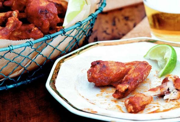 A basket of Sriracha chicken wings, two glasses of beer, and a plate with one eaten and one uneaten chicken wing and a wedge of lime.