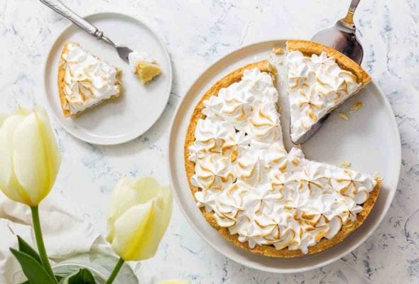 A lemon meringue tart in a white dish, with one slice on a plate beside it and one more being lifted out.