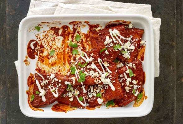 White casserole dish with cheese enchiladas, red sauce, sour cream, crumbled cotija cheese, and chopped cilantro