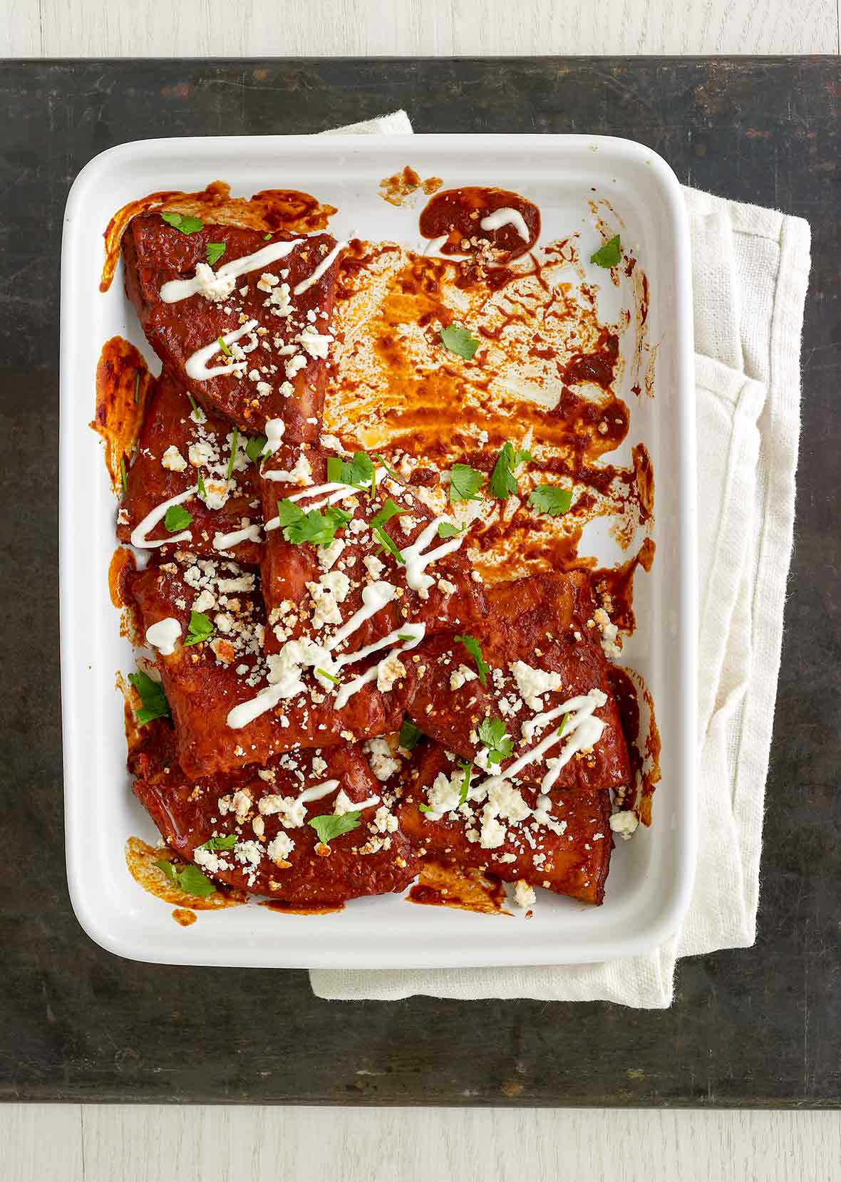 White casserole dish with cheese enchiladas, red sauce, sour cream, crumbled cotija cheese, and chopped cilantro