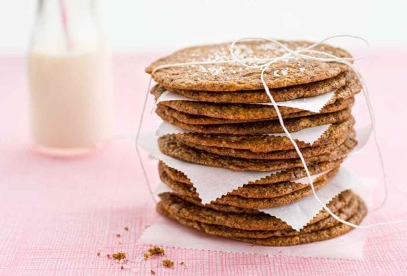 A stack of twisted Toll house cookies with pieces of parchment in between, tied up with string and a glass bottle of milk in the background.