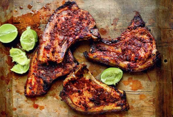 Four adobo marinated pork chops and several lime halves, some squeezed, on a wooden chopping block.
