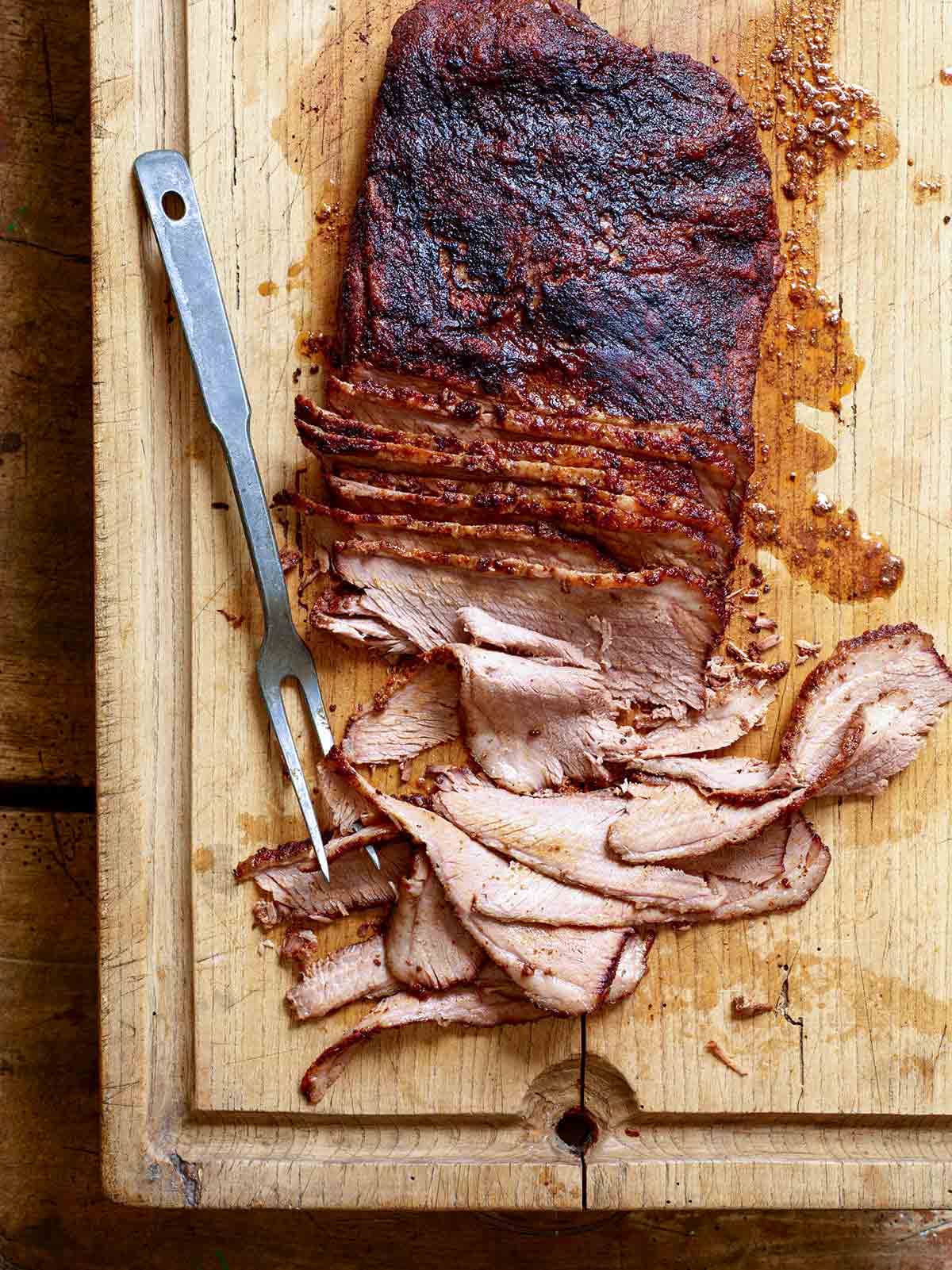 A partially sliced smoked brisket with coffee on a wooden cutting board with a meat fork resting beside it.