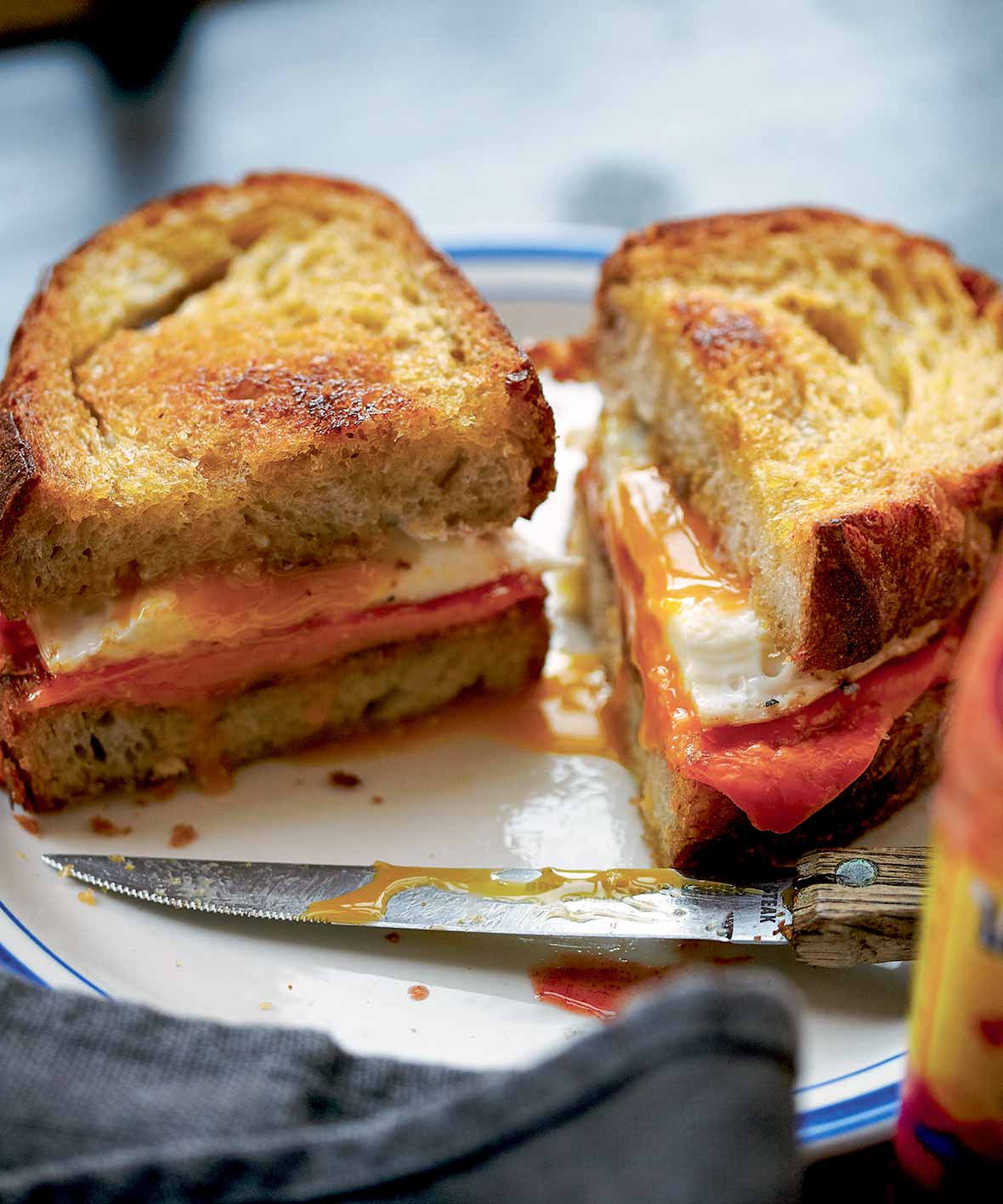 A fried egg sandwich cut in half on a white and blue plate with the knife resting in front.