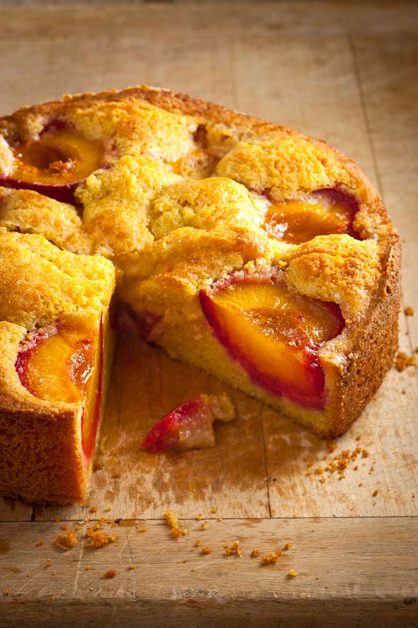 A round polenta plum cake on a wooden table with one slice cut from it.