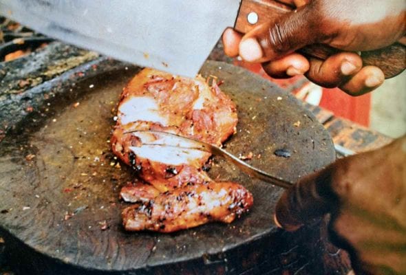 A sliced jerk chicken breast on a round cutting board, with a man's hand holding a cleaver above it