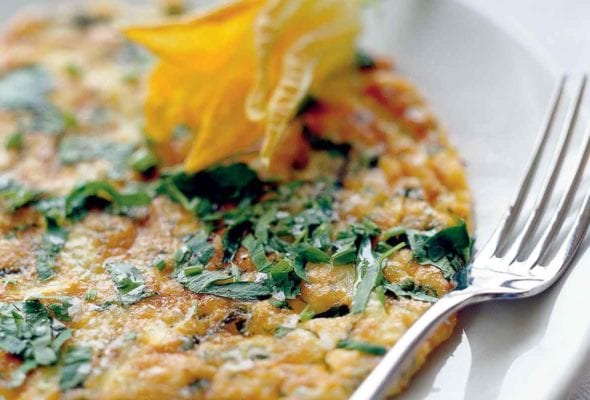 An Italian frittata garnished with a squash blossom on a white plate with a fork resting beside it.