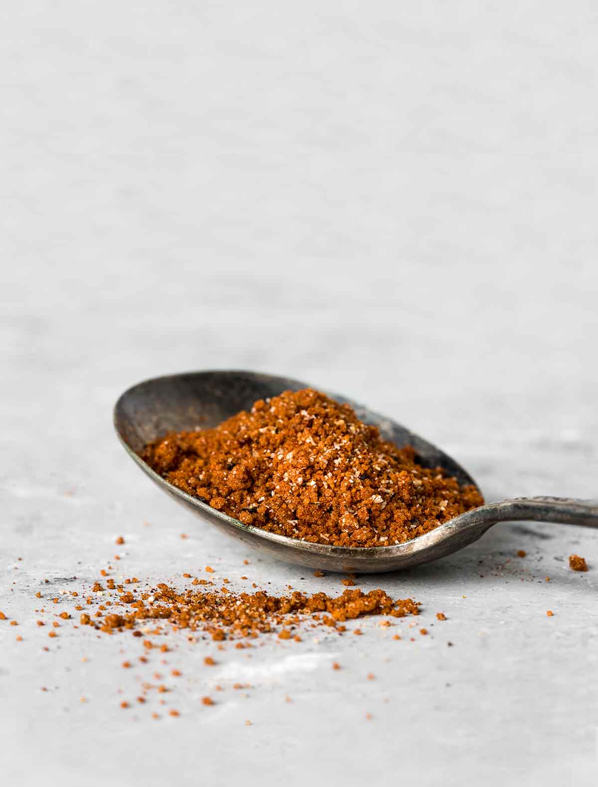 A spoon filled with Moroccan spice rub on a white surface with a little sprinkled beside the spoon.