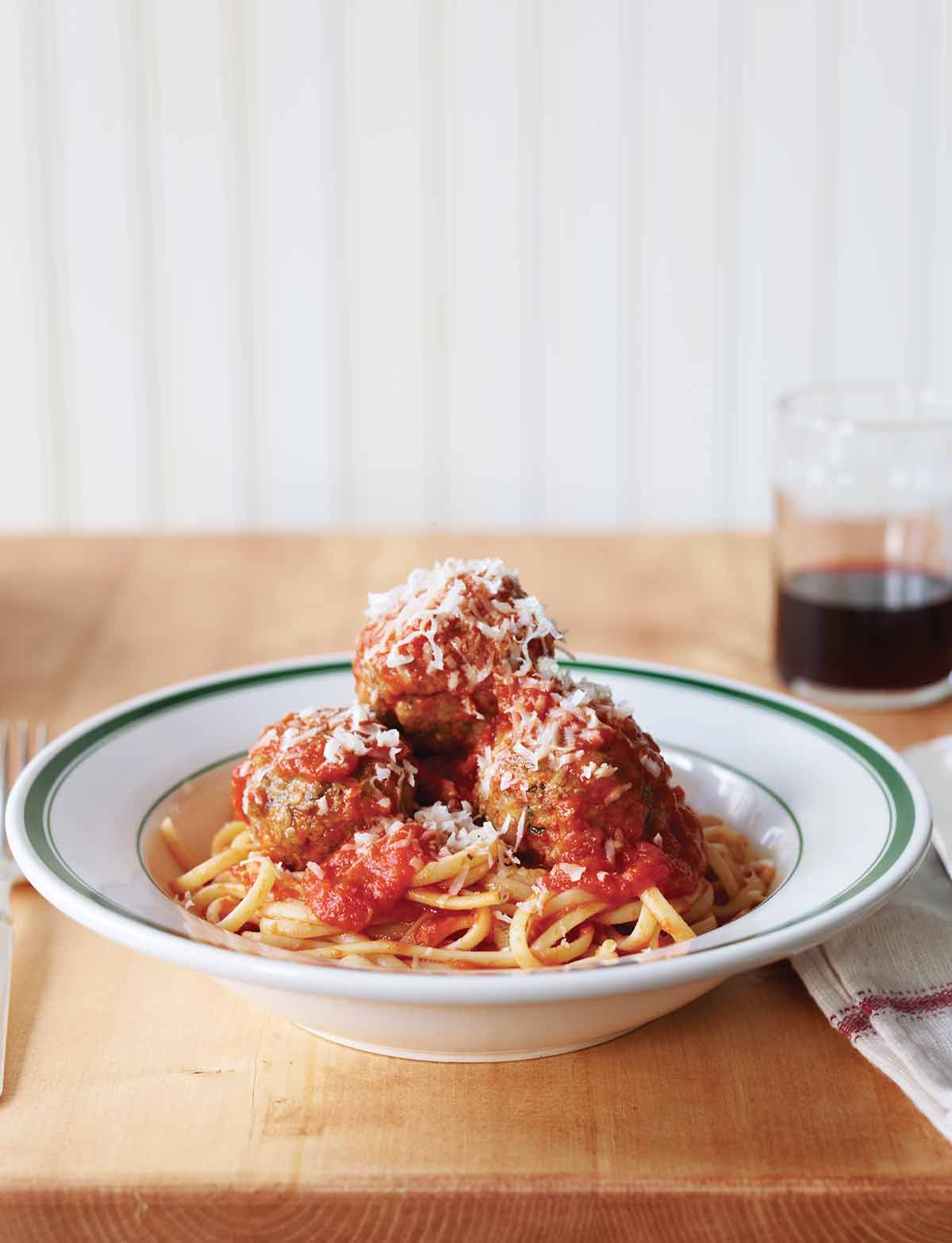 Three classic beef meatballs piled on pasta in a white and green bowl an finished with Parmesan.