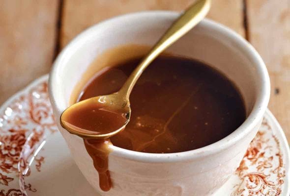 Easy caramel sauce in a teacup on a patterned saucer with a gold spoon resting on top and some caramel dripping down the side of the cup.