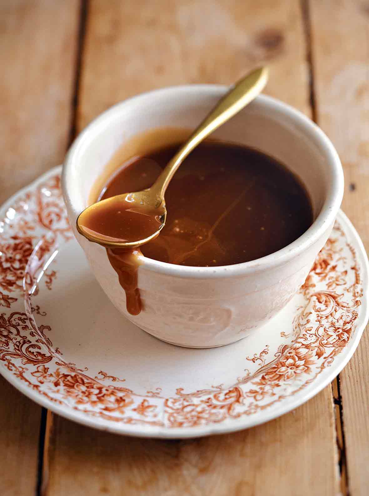 Easy caramel sauce in a teacup on a patterned saucer with a gold spoon resting on top and some caramel dripping down the side of the cup.