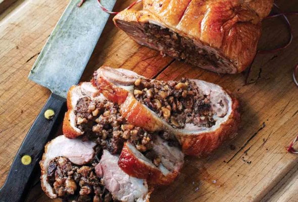 A roast duck stuffed with farro, figs, and hazelnuts, with three slices cut from it on a wooden cutting board with a knife resting beside it.