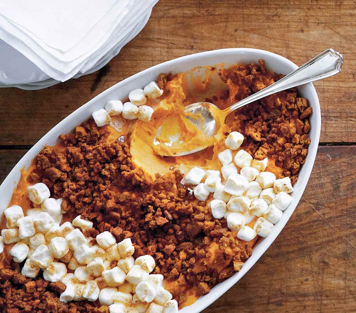 A white casserole dish filled with gingersnap and sweet potato casserole, topped with mini marshmallows and a spoon resting inside.