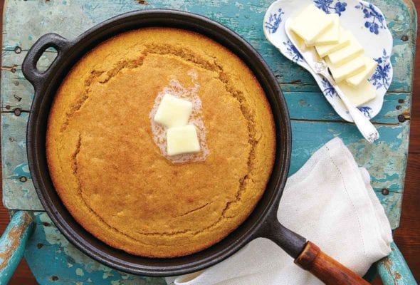 A cast iron skillet filled with cooked sweet potato cornbread topped with two pats of butter, resting on a old blue wooden chair with a dish of butter beside it.