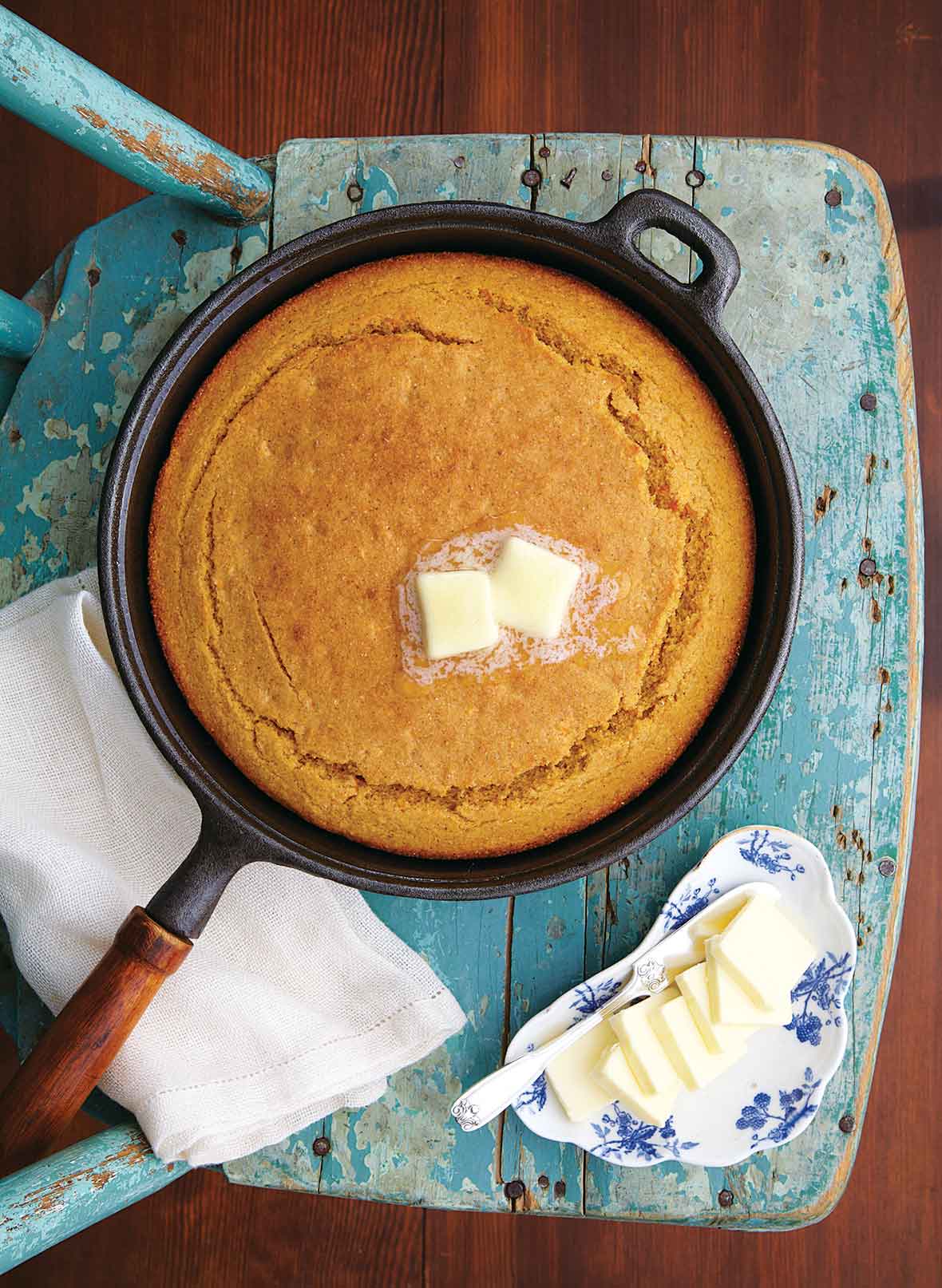 A cast iron skillet filled with cooked sweet potato cornbread topped with two pats of butter, resting on a old blue wooden chair with a dish of butter beside it.