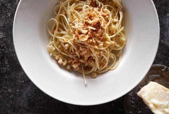 A white bowl filled with spaghetti topped with bread crumbs and a chunk of pecorino beside the bowl.