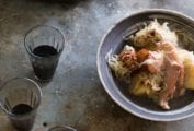 A Dutch oven and a porcelain bowl filled with choucroute garnie, made with pork, sausage, and sauerkraut, with two glasses of wine on the side.