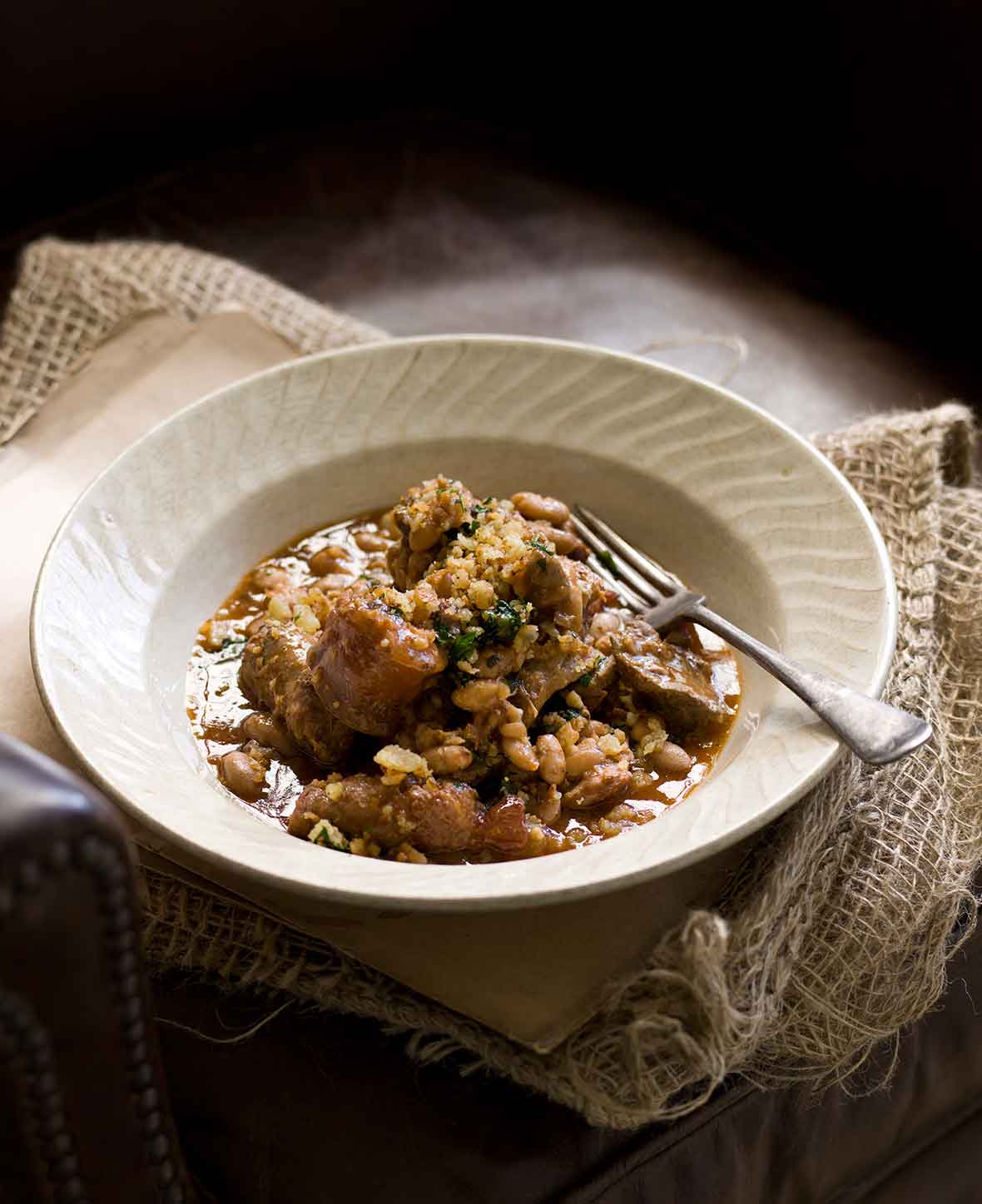 A white porcelain bowl filled with cassoulet and a fork rests on the side