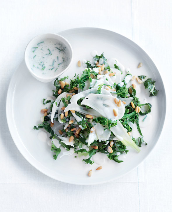 A white plate of kale slaw with shaved fennel and toasted pine nuts on top and a bowl of buttermilk dressing on the side.