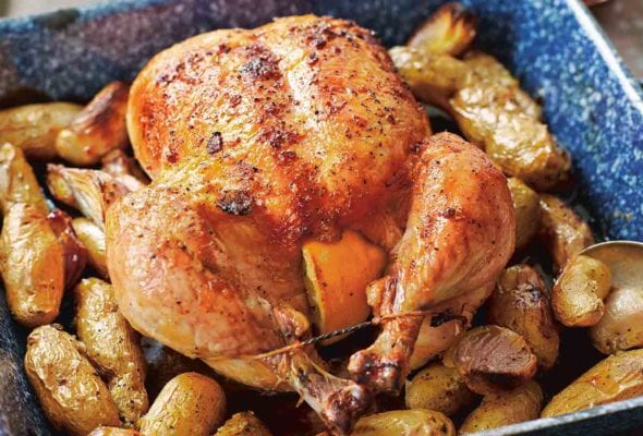 A whole maple roast chicken set on top of potatoes and shallots in a blue baking dish.