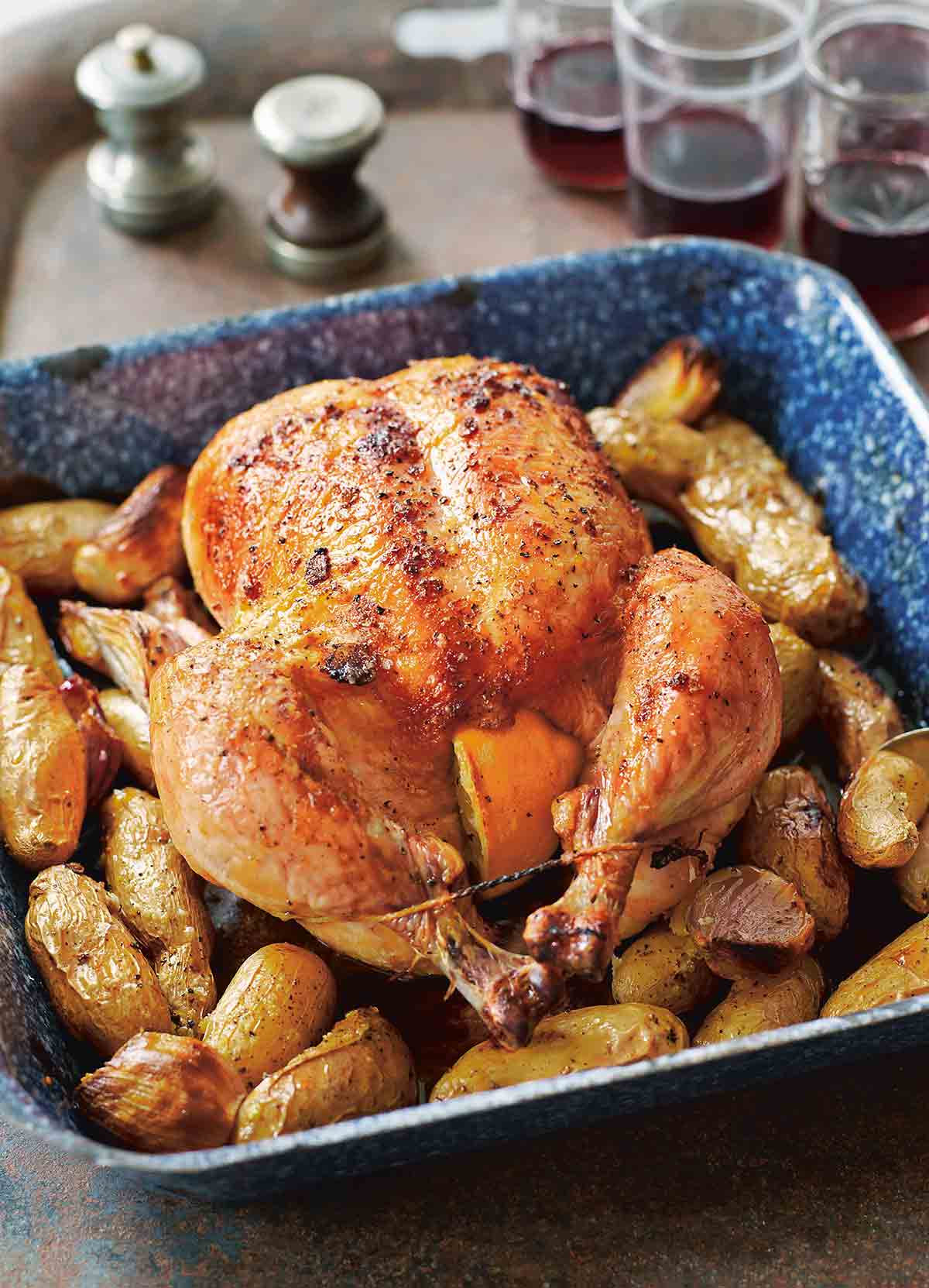 A whole maple roast chicken set on top of potatoes and shallots in a blue baking dish.
