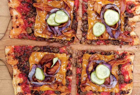 A bacon cheeseburger pizza topped with beef, bacon, pickles, cheese, and red onion, cut into four squares on a wooden pizza peel.